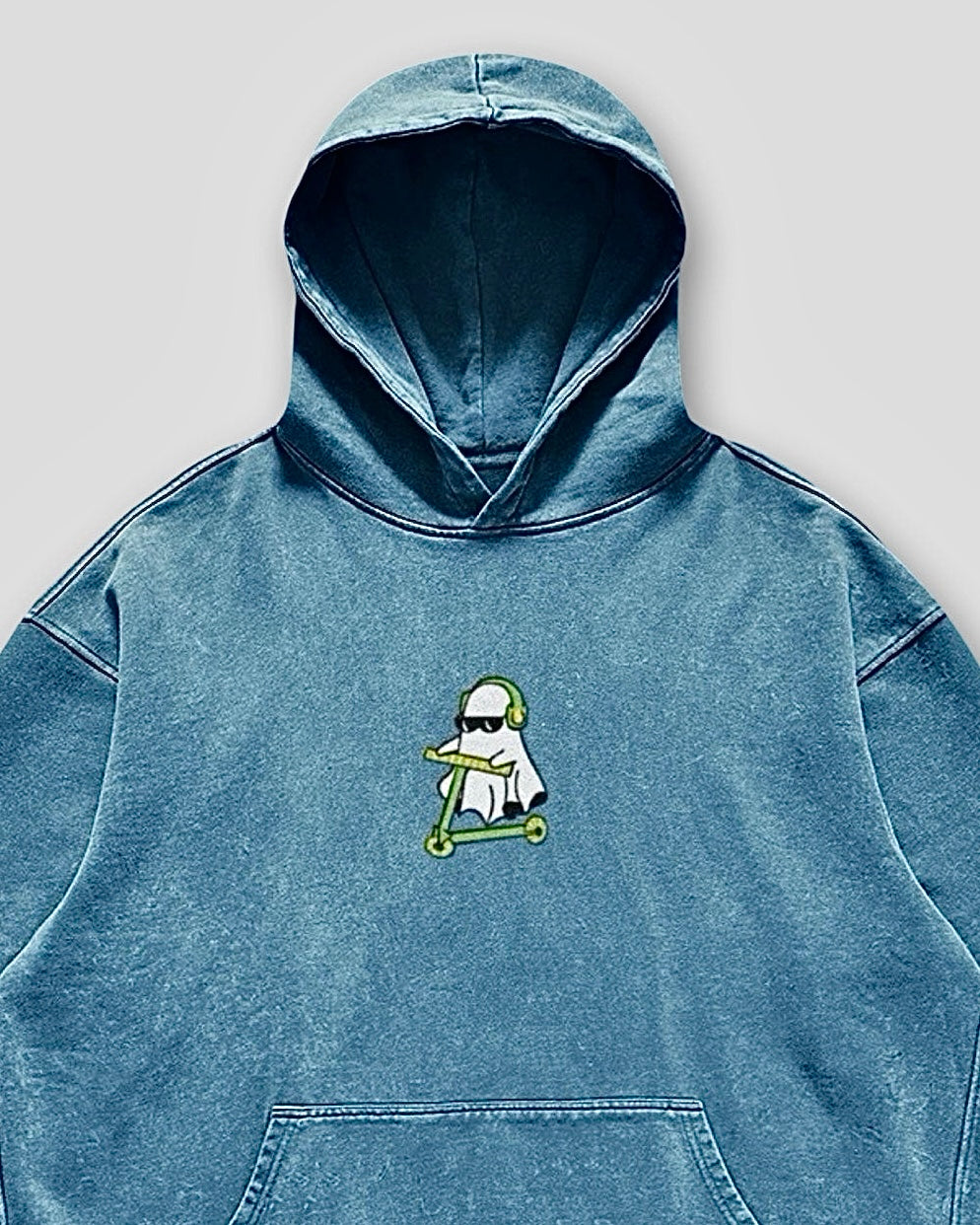 details view of a denim blue vintage washed heavyweight cotton oversized hoodie with a kangaroo pocked and embroidered original ghost graphics on the front and left arm against a white background