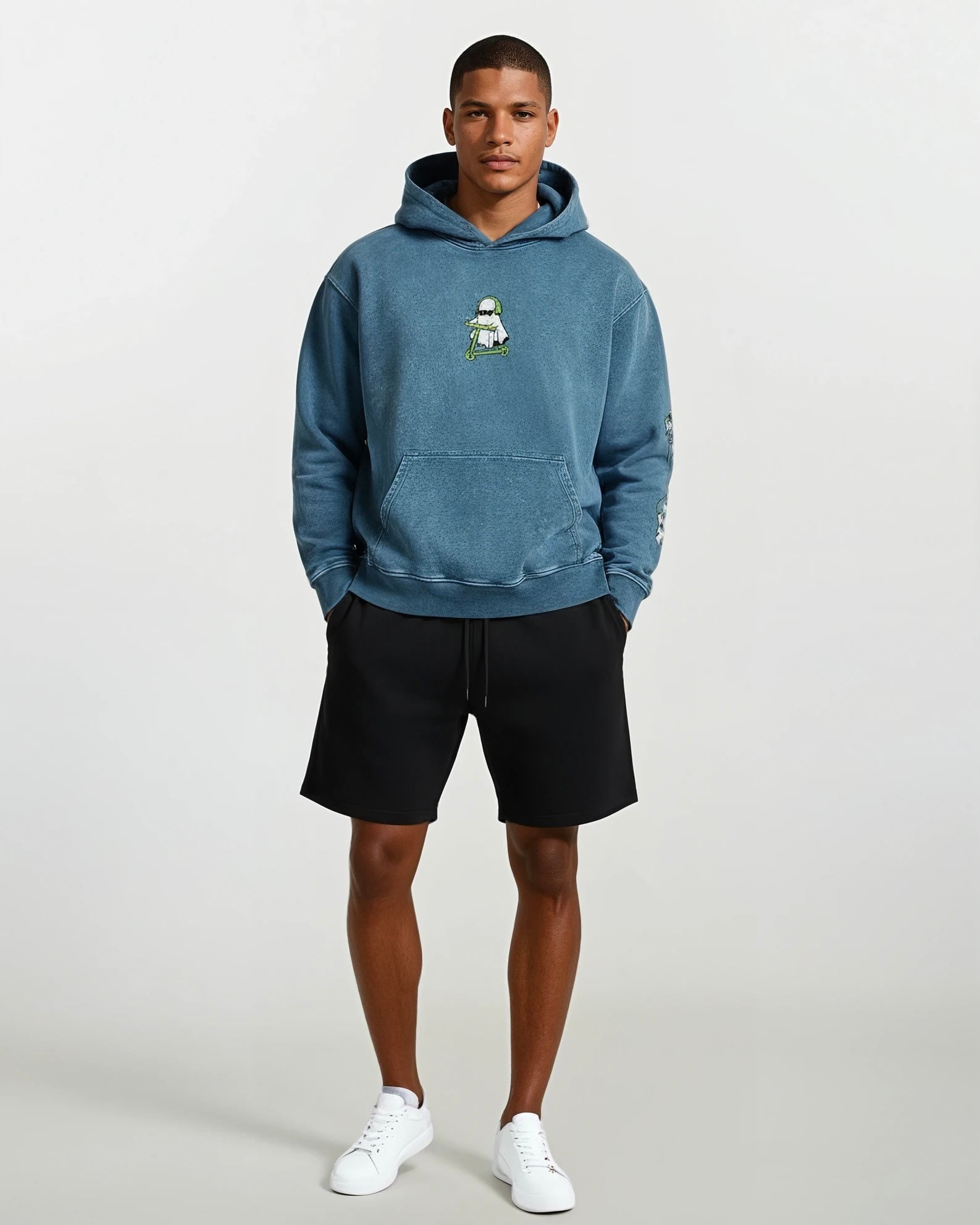 man wearing a denim blue vintage washed heavyweight cotton oversized hoodie with a kangaroo pocked and embroidered original ghost graphics on the front and left arm against a white background