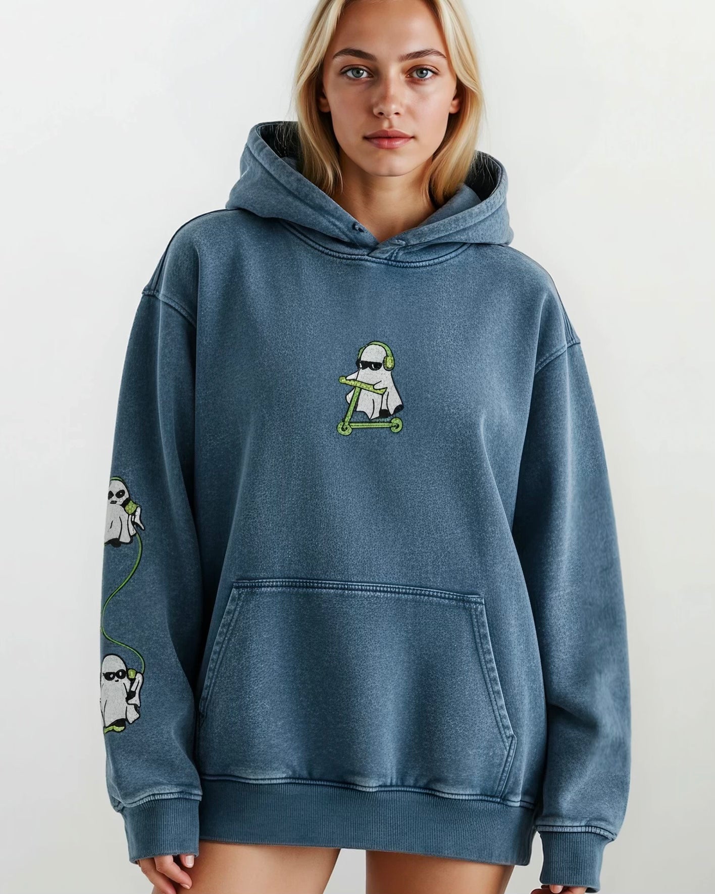 woman wearing a denim blue vintage washed heavyweight cotton oversized hoodie with a kangaroo pocked and embroidered original ghost graphics on the front and left arm against a white background