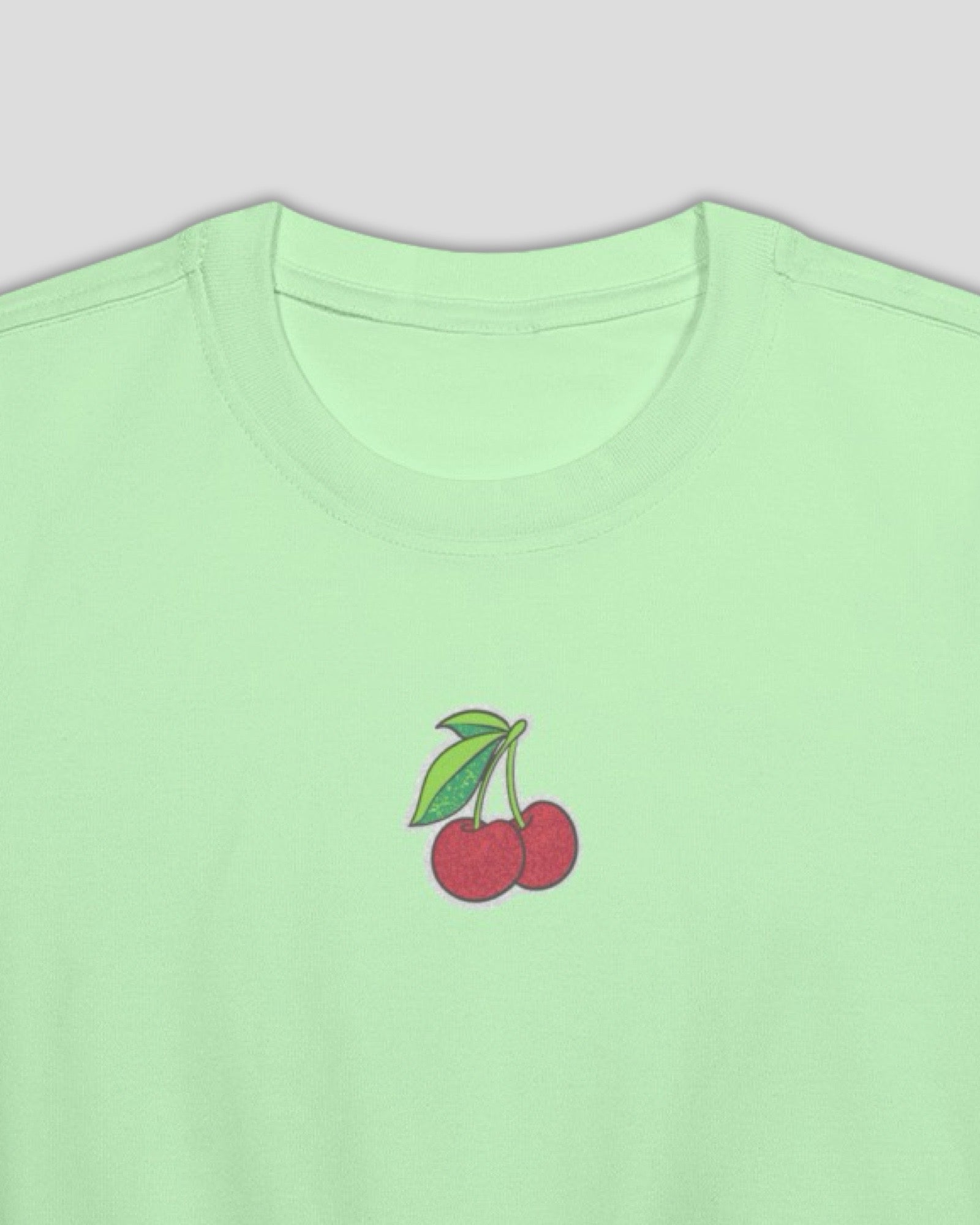 a close up shot of a green cropped cotton t-shirt with a small cherry embroidered design against a white background. 