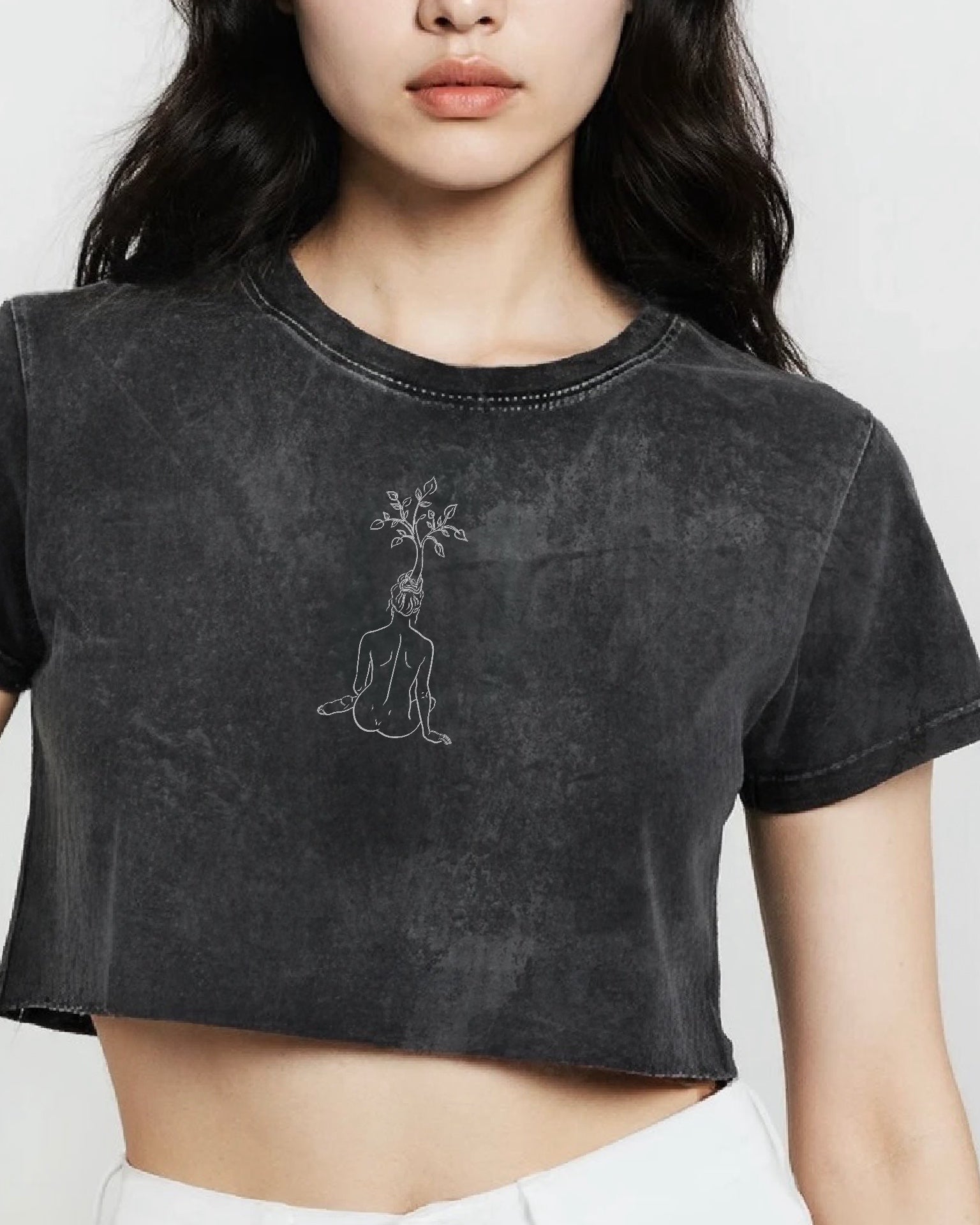 Person wearing a black crop top tee with a line design of a woman with a tree growing out of her head. photographed against a white background