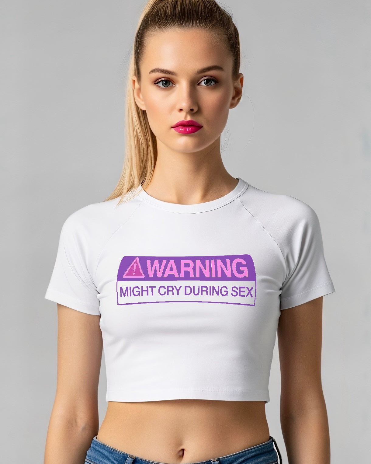 Person wearing a white crop baby tee with a purple warning label on a gray background